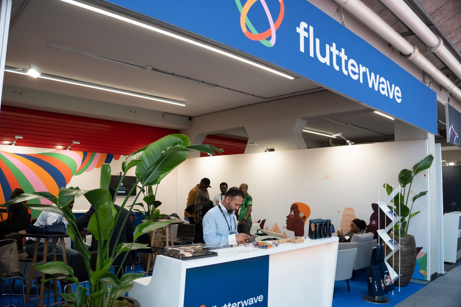 A bright and modern exhibition stand for Flutterwave at Money 20/20 Europe 2025. A team member is assisting a guest at the white reception desk, which features the Flutterwave logo. The booth has prominent blue branding, comfortable seating areas for guests, and is decorated with large green plants.