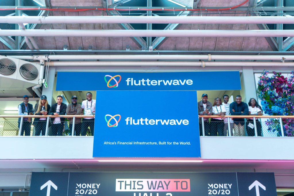 The Flutterwave delegation standing on a balcony over large blue Flutterwave corporate banners at the Money 20/20 Europe 2026 event hall in Amsterdam.